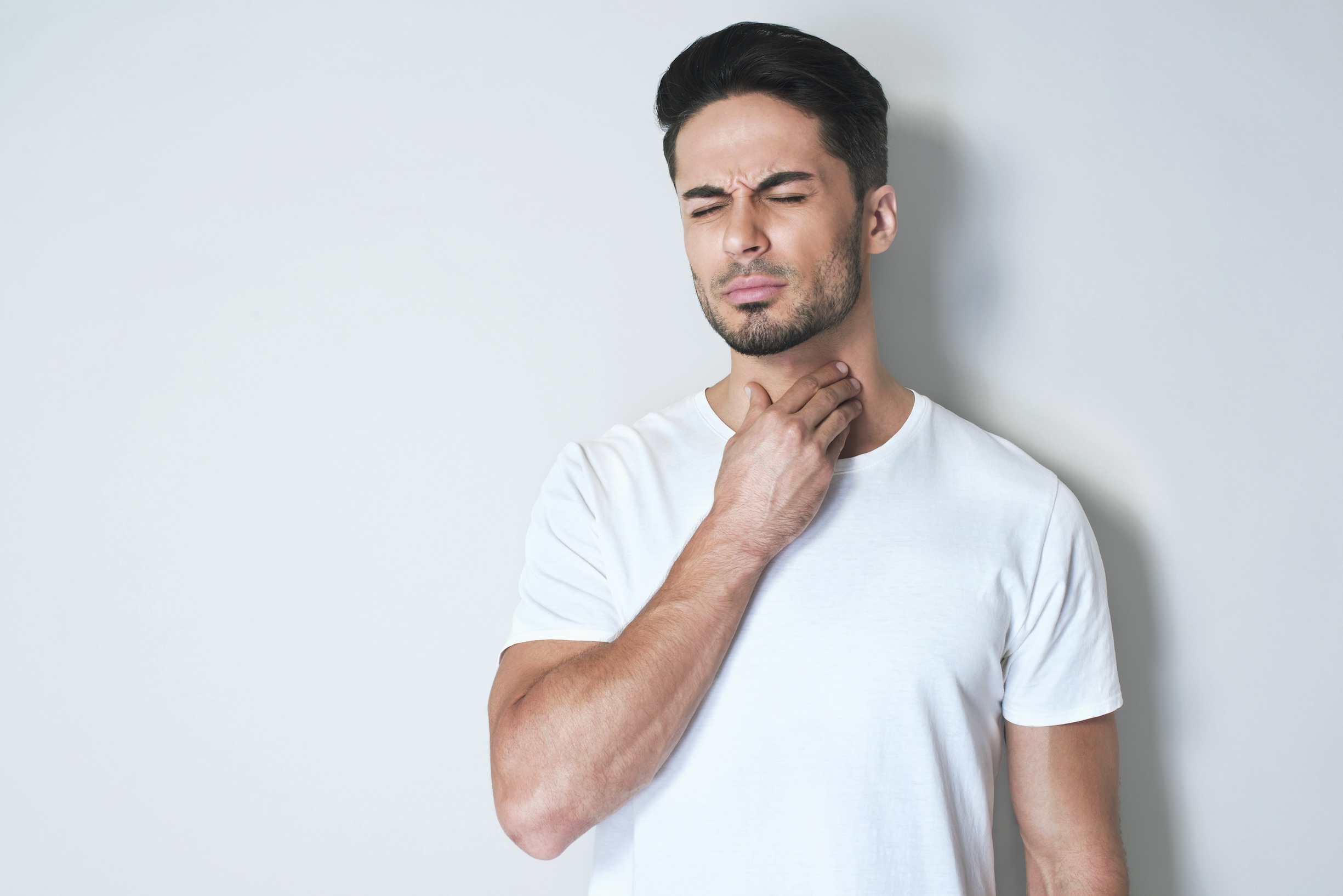 Young man having sore throat and touching his neck, wearing a loose white t-shirt against light grey background. Having difficulty swallowing.