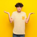 Young man over isolated yellow wall while raising hands, questioning where exactly semen is stored.